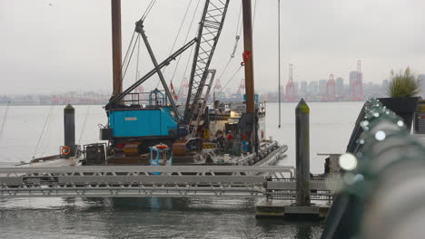 Arbeiter,-Kaum-Sichtbar,-Arbeiten-Auf-Einem-Mit-Einem-Kran-Geschmückten-Handelsboot,-Das-Auf-Dem-Wasser-Schwimmt,-Mit-Der-Skyline-Von-Vancouver-Am-Fernen-Horizont