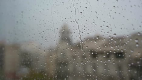 View-of-raindrops-landing-on-a-window-glass-during-heavy-rainfall,-,-with-an-urban-city-landscape-in-the-background