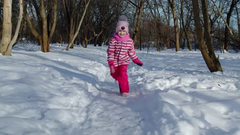 Niña-Alegre-Corriendo-Hacia-Un-Camino-Nevado,-Mirando-La-Cámara-En-El-Bosque-Del-Parque-De-Invierno