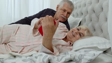 Pareja-De-Ancianos-En-Pijama-Acostados-En-La-Cama-Mirando-El-Teléfono-Móvil-Riendo-Y-Divirtiéndose