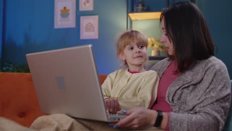 Madre-E-Hija-Pequeña-Estudian-En-Una-Computadora-Portátil,-Ven-Películas-Divertidas,-Se-Relajan,-Se-Tumban-En-El-Sofá