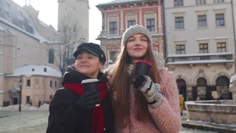 Two-smiling-women-tourists-traveling-together,-drinking-hot-tea,-coffee-from-cups-on-city-street