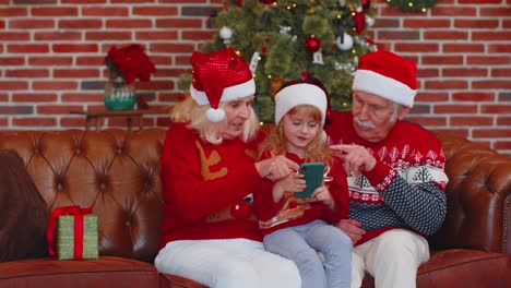 Abuelos-De-La-Familia-Mayor,-Nieta-Compran-Regalos-De-Navidad-En-Línea-En-Teléfonos-Móviles,-Compras