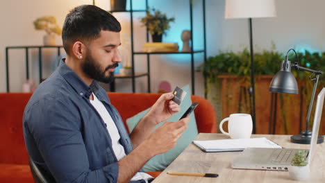 Indian-man-freelancer-using-credit-bank-card-and-smartphone-while-transferring-money-at-home-office