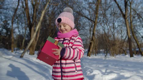 Alegre-Niña-Sonriente-Mirando-La-Cámara-Con-Caja-De-Regalo-De-Navidad-En-El-Parque-Nevado-De-Invierno