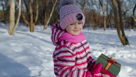 Niño-Sonriente-En-Un-Parque-Forestal-Nevado-De-Invierno-Mirando-La-Cámara,-Sosteniendo-Una-Caja-De-Regalo-De-Navidad