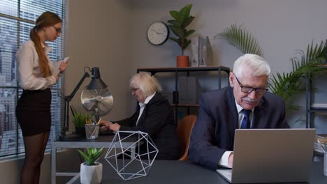 Cheerful-mature-old-businessman-raising-hands-celebrating-sudden-victory-with-colleagues-in-office