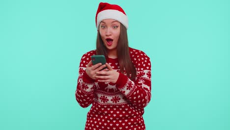 Una-Joven-Adolescente-Con-Un-Suéter-Navideño-Mirando-La-Pantalla-Del-Teléfono-Inteligente-Regocijándose-Sinceramente-Por-Ganar-éxito-Y-Suerte