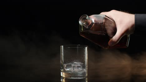 Barman-pour-whiskey-cognac-brandy-from-bottle-into-drinking-glass-with-ice-cubes-on-dark-background