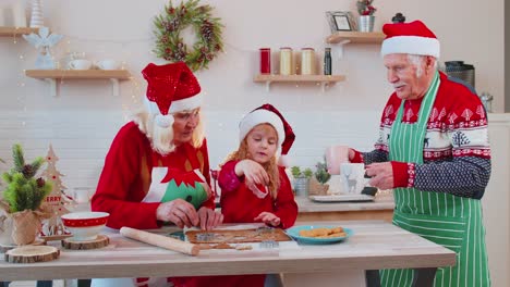 Senior-man-with-cups-hot-chocolate-walking-into-Christmas-home-kitchen-to-grandmother-and-grandchild