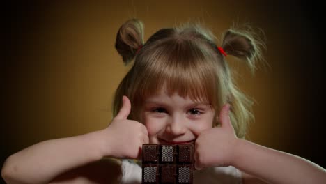 Niña-Pequeña-Comiendo-Postre-De-Barra-De-Chocolate,-Mostrando-Los-Pulgares-Hacia-Arriba-Aislados-Sobre-Fondo-Oscuro