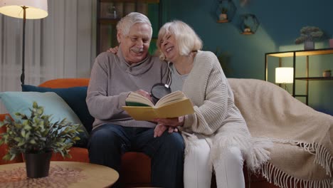 Senior-grandparents-couple-relaxing,-reading-book,-talking-enjoying-leisure-hobbies-at-night-home