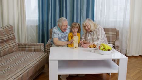 Pareja-De-Ancianos,-Abuelos-Y-Nieta-Disfrutando-De-Un-Juego-De-Mesa-Construyendo-Una-Torre-A-Partir-De-Bloques-En-Casa