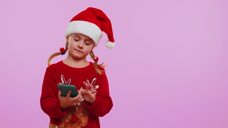 Niña-Pequeña-Con-Sombrero-De-Papá-Noel-Navideño-Con-Teléfono-Móvil-Mostrando-Un-Lugar-Vacío,-área-De-Publicidad