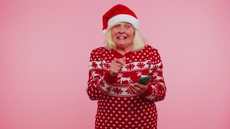 Abuela-En-Suéter-De-Navidad-Mirando-La-Pantalla-Del-Teléfono-Inteligente-Regocijándose-Sinceramente-Ganar-éxito-Suerte