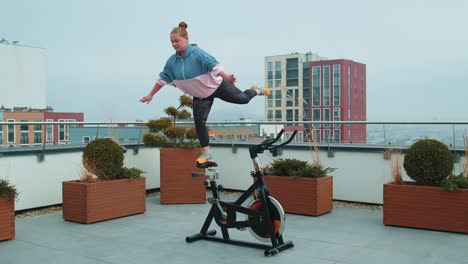 Chica-Atlética-Realizando-Ejercicios-De-Trucos-Acrobáticos-De-Golondrina-En-Bicicleta-Estática-En-El-Techo-De-La-Casa