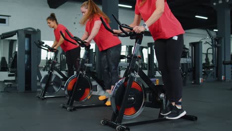 Athletic-women-group-riding-on-spinning-stationary-bike-training-routine-in-gym,-weight-loss-indoors