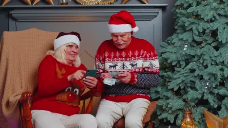 Pareja-De-Familiares-Mayores-Buscando-Comprar-Regalos-De-Navidad-En-Línea-En-Una-Aplicación-De-Teléfono-Móvil-En-Casa