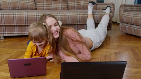 Una-Niñera-Y-Una-Niña-Estudiando-Junto-Con-Una-Computadora-Portátil,-Mientras-Están-Acostadas-En-Un-Piso-Cálido-En-Casa