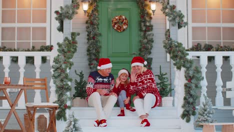 Abuela-Mayor-Abuelo-Con-Nieta-Sentada-En-El-Porche-De-La-Casa-De-Navidad-Saludando-Hola