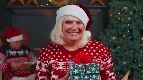 Senior-grandmother-in-festive-sweater-presenting-Christmas-gift-box,-smiling,-looking-at-camera