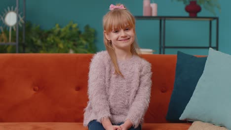 Portrait-of-little-adorable-child-girl-sitting-alone-on-sofa-at-home-looking-at-camera-and-smiling