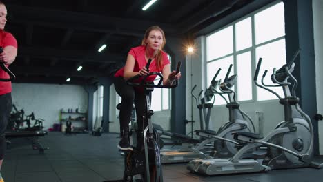 Gruppenunterricht-Für-Zwei-Befreundete-Frauen,-Die-Im-Modernen-Fitnessstudio-Auf-Einem-Heimtrainer-Trainieren-Und-Spinning-Machen