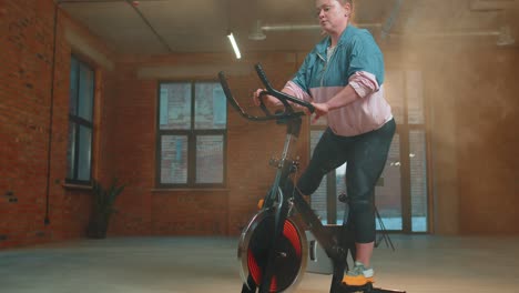 Mujer-Atlética-Montando-Una-Rutina-De-Entrenamiento-En-Bicicleta-Estacionaria-Giratoria-En-Un-Gimnasio-De-Niebla,-Pérdida-De-Peso-En-Interiores