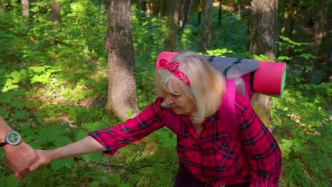 Elderly-woman-on-hikinig-adventure-trip-holding-mans-hand-helping-her-climb-on-mountain-hill-in-wood