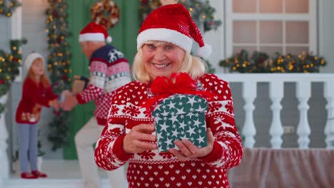 Portrait-of-grandmother-woman-presenting-gift-box-smiling-near-decorated-Christmas-house-with-family