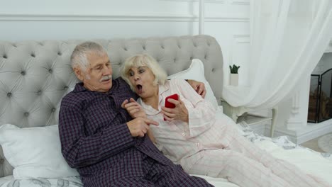 Pareja-De-Ancianos-En-Pijama-Acostados-En-La-Cama-Mirando-Por-Teléfono-Móvil-Haciendo-Compras-En-Línea