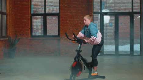 Chica-Atlética-Realizando-Ejercicios-De-Entrenamiento-Aeróbico-En-Bicicleta-Estática-En-Un-Gimnasio-Con-Niebla