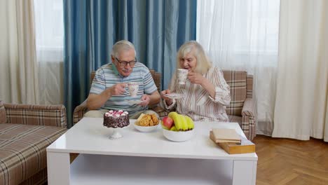 Pareja-Mayor-Sosteniendo-Té-Hablando,-Riendo,-Sonriendo,-Besándose,-Sentado-En-El-Sofá-En-Casa