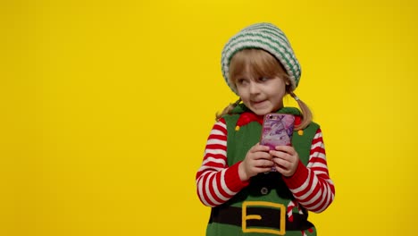 Una-Niña-Con-Un-Teléfono-Móvil-Disfrazada-De-Elfo-Navideño-De-Ayudante-De-Papá-Noel-Señala-Con-El-Dedo-A-Un-Lado-En-Una-Pared-En-Blanco