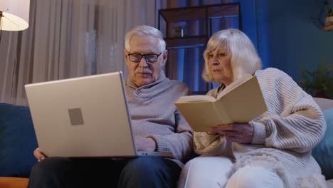 Senior-grandparents-couple-reading-book,-using-laptop-pc-on-couch-in-night-living-room-at-home