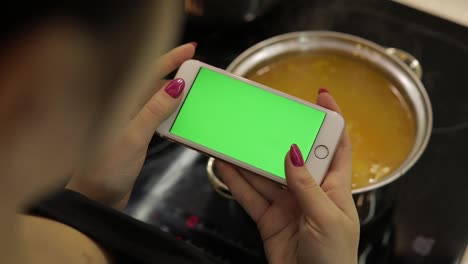 Woman-is-holding-a-smartphone-with-a-green-screen,-and-cooking-soup