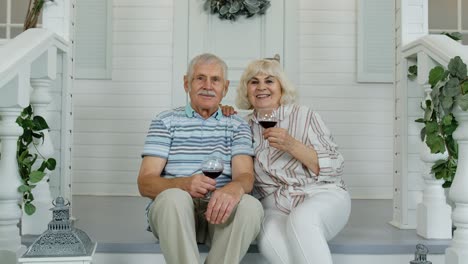 Attractive-senior-elderly-Caucasian-couple-sitting-and-drinking-wine-in-porch-at-home,-making-a-kiss