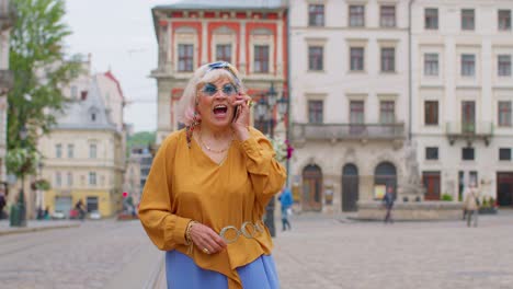 Senior-old-tourist-woman-in-stylish-clothes-talking-on-mobile-phone-while-walking-on-city-street