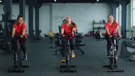 Group-of-smiling-friends-women-class-exercising,-training,-spinning-on-stationary-bike-at-modern-gym