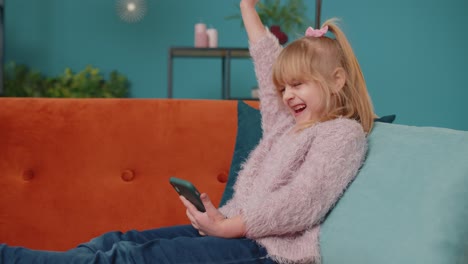 Una-Niña-Feliz-Y-Pequeña-Usa-Un-Teléfono-Móvil-Haciendo-Un-Gesto-Ganador-Y-Celebra-Sola-En-El-Sofá-De-Casa