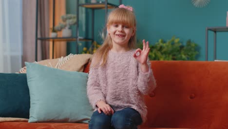 Portrait-of-little-child-girl-kid-alone-on-sofa-at-home-showing-ok-gesture,-like-sign,-positive-good