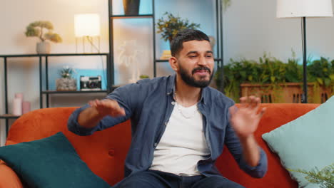 Energetic-happy-Indian-man-singing-dancing-with-hands-listening-music-sitting-on-couch-in-home-room