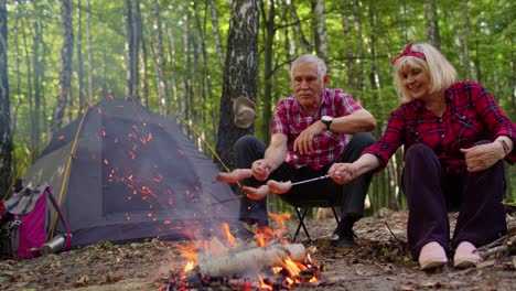 Happy-senior-man-and-woman-talking,-cooking-frying-sausages-over-campfire-in-evening-wood,-camping