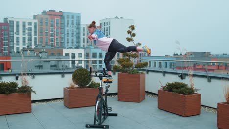 Athletic-girl-performing-aerobic-riding-training-exercises-on-cycling-stationary-bike-on-house-roof