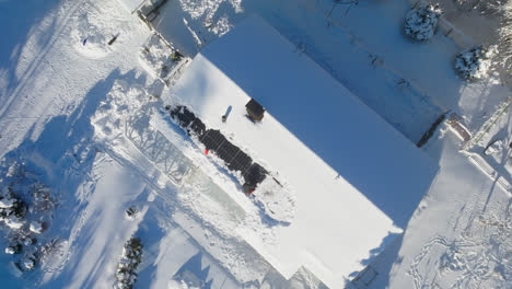 Aerial-view-rising-above-a-man-clearing-snow-from-a-solar-cell-roof,-winter-day