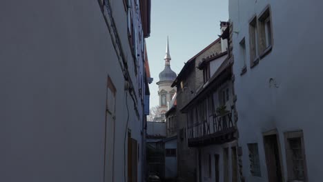 Birds-flying-over-small-gobblestone-road-with-half-timbered-houses,-Church-seen-through-the-beautiful-buildings