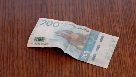 Mano-Colocando-Un-Billete-Noruego-De-200-Kr-En-El-Centro-De-Una-Mesa-De-Madera