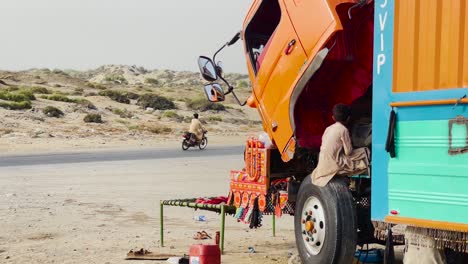 Static-shot-of-a-boy-helping-repair-engine-of-a-container-truck-by-the-side-of-a-road-after-it-broke-down-beside-a-highway-at-daytime