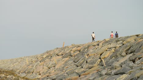 Young-men-walk-on-high-barrier-of-rocks-by-harbor-at-Cape-Coast,-Ghana