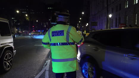 Nachts-Steht-Ein-Polizeibeamter-Einer-Großstadt-An-Einer-Kreuzung-Am-Parliament-Square-Und-Fährt-Mit-Seinem-Fahrzeug-Weg,-Während-Ein-Alter-Schwarzer-Londoner-Bus-Vorbeifährt-Und-Eine-Abgaswolke-Ausstößt